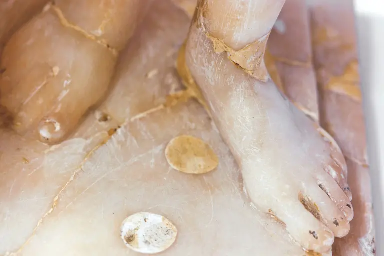 Das Bild zeigt eine detaillierte Nahaufnahme eines Fußes, der aus Marmor oder Alabaster gefertigt ist. Der Fuß ist unvollständig und weist einige Risse auf. Der Untergrund ist ebenfalls sichtbar, einschließlich runder Elemente, die darauf liegen.
