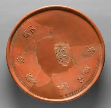 Runde, terrakottafarbene Schale mit eingravierten Tiermotiven. Teile der Schale sind beschädigt. Die Motive zeigen stilisierte Darstellungen von verschiedenen Tieren, die gleichmäßig um den Rand angeordnet sind. Die Oberfläche ist glatt und der Farbton ist einheitlich rotbraun.