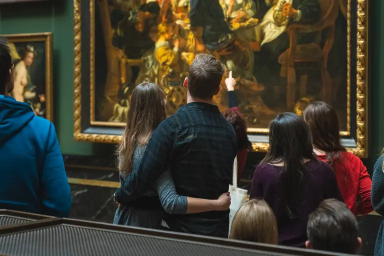 Eine Gruppe von Besuchern in einem Museum steht vor einem großen Gemälde. Einige Personen zeigen auf das Kunstwerk, während sie es aufmerksam betrachten. Der Raum ist elegant gestaltet, mit üppigen Rahmen um die Kunstwerke.