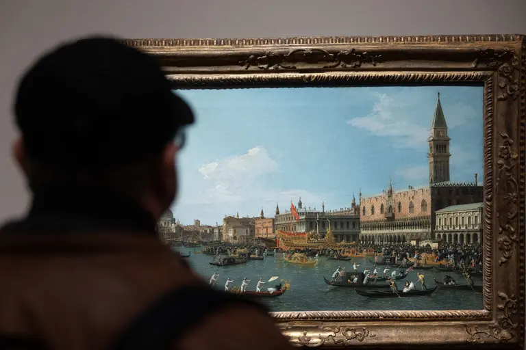 A person in a brown jacket and black hat is viewing a large painting of a Venetian scene, featuring boats on the canal and historic buildings under a blue sky. The painting is framed in an ornate gold frame.