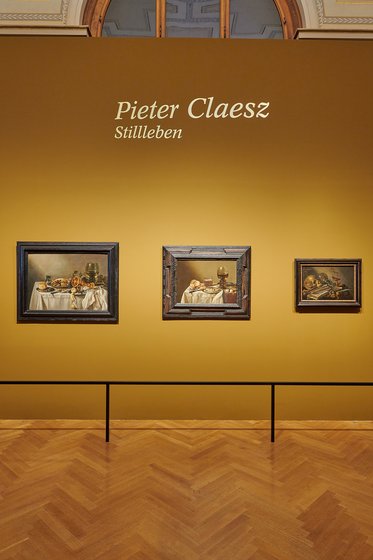 Ein Ausstellungsraum mit einer gelben Wand, auf der der Schriftzug „Pieter Claesz Stillleben“ zu sehen ist. Drei Stillleben-Gemälde sind in schwarzen Rahmen an der Wand angebracht. Der Boden ist aus Holz.
