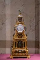 Eine kunstvoll gestaltete goldene Standuhr mit einem großen Zifferblatt und bunten Verzierungselementen. Die Uhr hat einen reich verzierten Sockel und eine Kuppel, die von kleinen Ornamenten geschmückt ist. Sie steht auf einem einfarbigen Untergrund in einem eleganten Raum.