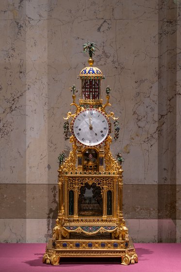 Eine kunstvoll gestaltete goldene Standuhr mit einem großen Zifferblatt und bunten Verzierungselementen. Die Uhr hat einen reich verzierten Sockel und eine Kuppel, die von kleinen Ornamenten geschmückt ist. Sie steht auf einem einfarbigen Untergrund in einem eleganten Raum.