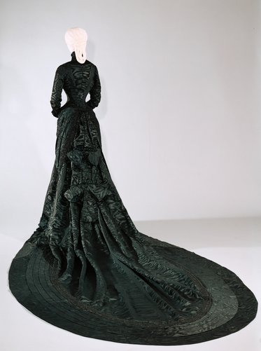 Ein elegantes, langes, schwarzes Kleid mit reich strukturiertem Stoff, das hinter einer schmalen Figur drapiert ist. Der Rock entfaltet sich in sanften Wellen und reicht bis zum Boden. Die Rückansicht zeigt die detaillierte Textur des Kleides, während die Figur mit geschlossenen Händen vor dem Körper steht.