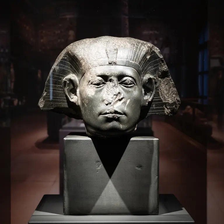 Eine antike Pharaonenbüste aus Stein, mit abgebrochenen Teilen am Gesicht und einem markanten Kopfschmuck. Die Skulptur steht auf einem schwarzen Sockel in einem museumähnlichen Raum mit schwachem Licht im Hintergrund.