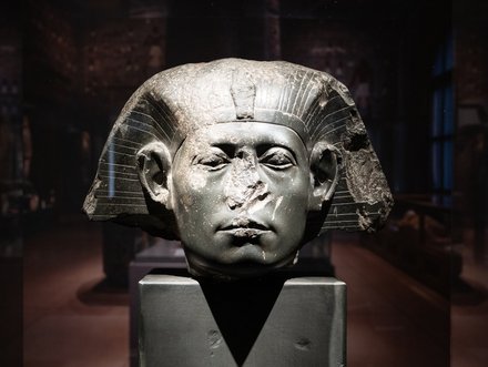 Eine antike Pharaonenbüste aus Stein, mit abgebrochenen Teilen am Gesicht und einem markanten Kopfschmuck. Die Skulptur steht auf einem schwarzen Sockel in einem museumähnlichen Raum mit schwachem Licht im Hintergrund.