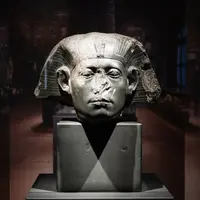Eine antike Pharaonenbüste aus Stein, mit abgebrochenen Teilen am Gesicht und einem markanten Kopfschmuck. Die Skulptur steht auf einem schwarzen Sockel in einem museumähnlichen Raum mit schwachem Licht im Hintergrund.