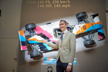 Ein Mann steht neben einem ausgestellten Rennwagen, der an der Wand montiert ist. Der Rennwagen ist bunt lackiert und trägt die Nummer 62. Oben sind technische Daten wie Leistung und Geschwindigkeit zu sehen. Der Mann lächelt und präsentiert den Wagen.