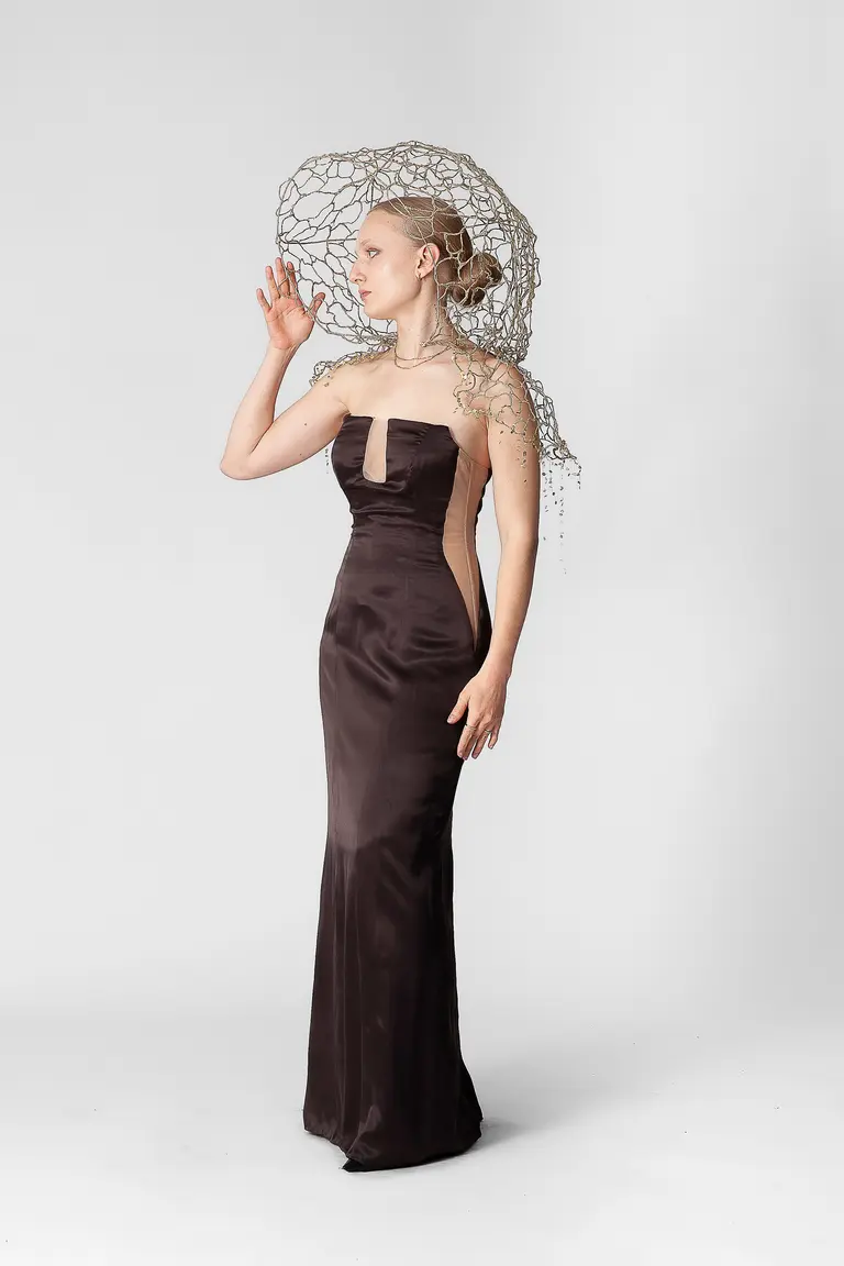 A model wears a form-fitting brown satin gown with a strapless design and a cutout detail. Her hairstyle features intricate braids and elaborate headpiece resembling a net. She poses gracefully against a neutral background, conveying elegance and artistry.