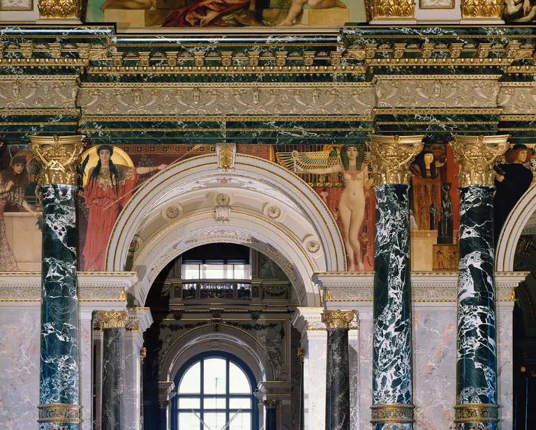 Ein kunstvoll dekoriertes Innenraum mit beeindruckenden fresken und Säulen. Die Wände zeigen historische Szenen und Figuren, während die Marmorsäulen eine majestätische Atmosphäre schaffen. Der Hintergrund deutet auf weitere architektonische Elemente hin, die zur Pracht des Raumes beitragen.