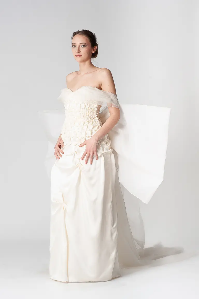 A model poses in an elegant, off-shoulder white gown adorned with floral embellishments. The dress features a flowing, translucent overlay and a long skirt. The background is plain to emphasize the garment. The model's expression is serene and confident.