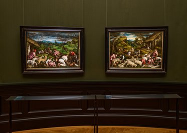 Zwei große Gemälde hängen an der Wand in einem Museum. Das linke Bild zeigt Menschen, die mit Tieren arbeiten, während das rechte Bild eine ländliche Szene mit weiteren Menschen und Tieren darstellt. Beide Bilder sind in für das Museum typischen Rahmen gefasst.