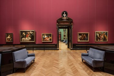 Ein Innenraum einer Kunstgalerie mit einem pinken Wande und mehreren Gemälden in schwarzen Rahmen. In der Mitte stehen zwei blaue Sofas. Im Hintergrund ist ein Zugang zu einem weiteren Raum sichtbar, der mit menschlichen Figuren bemalt ist.