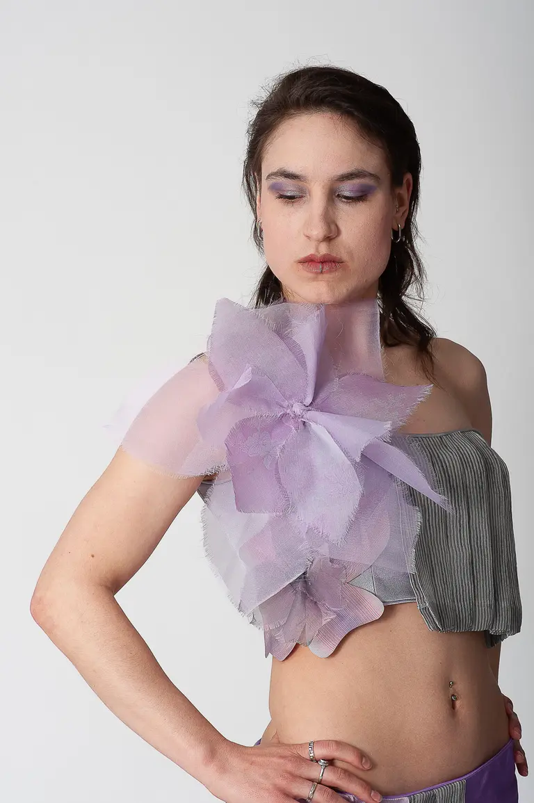 A person poses with a confident expression, wearing a unique, handmade crop top that features a large, translucent purple flower on one shoulder. The background is plain white, allowing the outfit to stand out. Their makeup includes soft purple tones, highlighting the creative style.