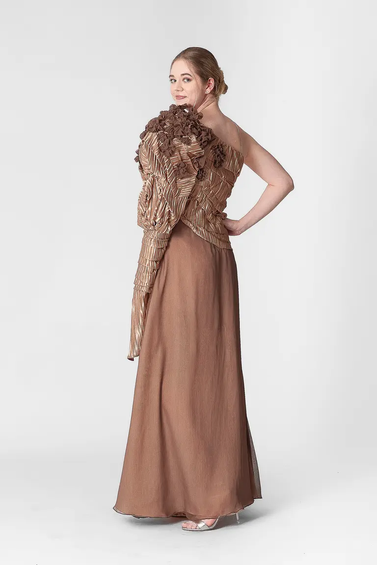 A woman poses in a unique, elegant one-shoulder dress adorned with textured fabric and floral accents. The dress is a warm brown hue, flowing to the floor. She has an updo hairstyle and is standing against a plain white background, showcasing the dress's design.