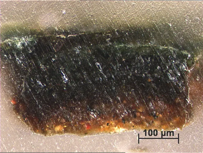 Mikroskopische Aufnahme eines Materials mit einer scharfen Kante. Die Textur zeigt verschiedene Farben, von Schwarz über Braun bis zu einem leicht grünen Farbton. Eine Maßstabsangabe von 100 Mikrometern ist im Bild sichtbar.