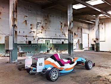 Ein stilisierter Rennwagen steht in einer verfallenen Industriehalle. Der Wagen hat ein buntes Design in den Farben Blau, Pink, Orange und Weiß und trägt die Nummer 62. Die Wände der Halle sind beschädigt und zeigen Spuren von Verfall.