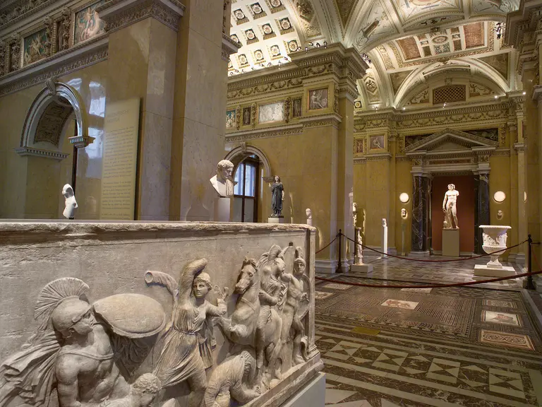 Eine weite Betrachtung eines Kunstmuseums mit antiken Skulpturen und reliefartigen Darstellungen auf einer Mauer. Im Hintergrund sind Statuen und Säulen in einem eindrucksvollen Raum mit kunstvoll gestalteter Decke zu sehen.