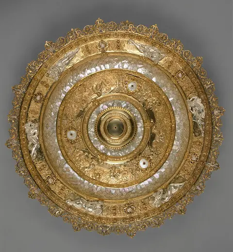 Detailreiche Deckenspiegel mit goldenen und perlmutternen Verzierungen, die spiralförmig angeordnet sind. In der Mitte befindet sich eine runde Vertiefung. Die äußeren Ränder sind mit filigranen Mustern und Figuren geschmückt. Der Hintergrund ist graufarben.