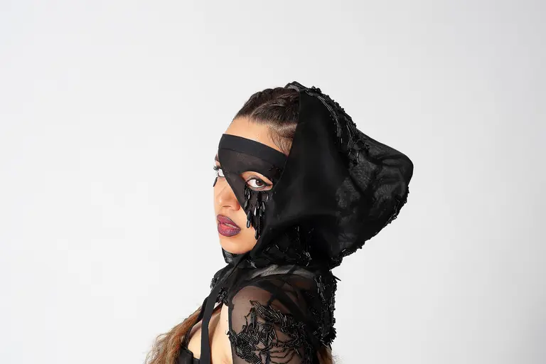 A woman poses with a dramatic expression, wearing a black, intricately designed headpiece that covers part of her face. She has long hair styled in loose waves and bold makeup, including dark lipstick. The background is plain white, emphasizing her striking look.