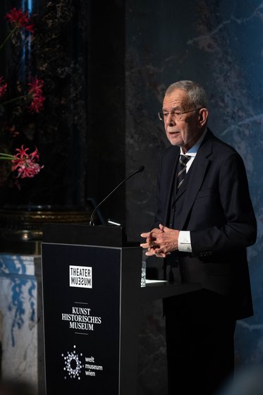 Ein älterer Mann steht am Rednerpult und hält eine Ansprache. Er trägt einen Anzug und eine Krawatte. Im Hintergrund sind Blumen und eine strukturierte Wand zu sehen.