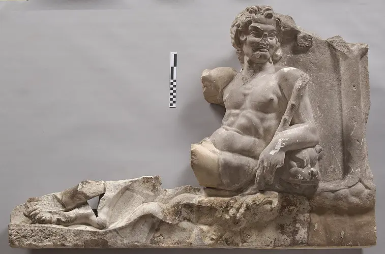 Eine antike Statue eines sitzenden männlichen Figures, teilweise beschädigt. Die Figur zeigt einen muskulösen Oberkörper, während sie mit einem Bein ausgestreckt und dem anderen angewinkelt sitzt. Die Darstellung umfasst auch Falten eines Gewandes, das über den Unterkörper drapiert ist.