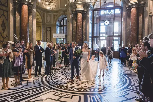 A bride and groom walk hand-in-hand through the festive foyer of the Kunsthistorisches Museum, surrounded by applauding guests. The bride wears a white gown and holds a bouquet, while a young girl follows, carrying the bride's train. The venue features ornate architecture and a patterned floor.