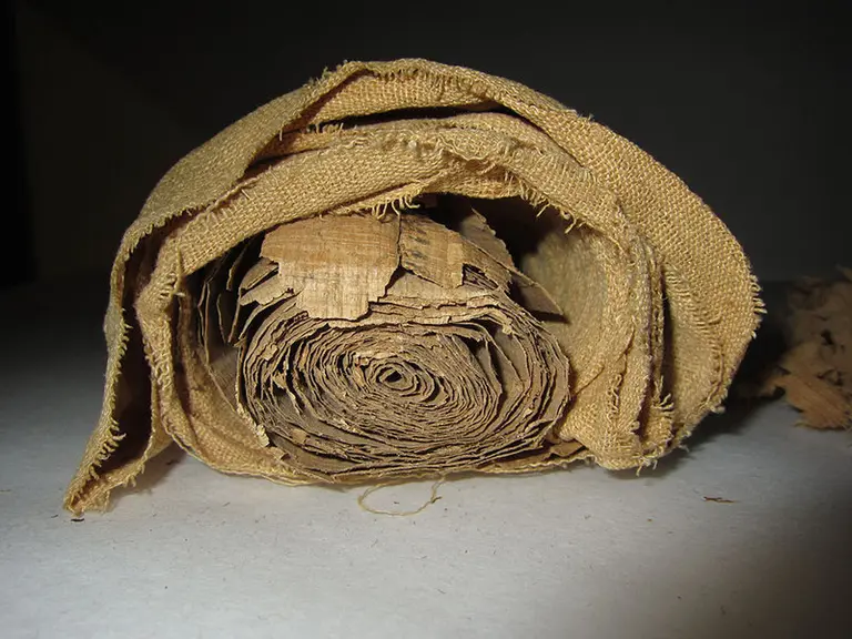 Eine gerollte, antike Schriftrolle aus Papier, die in einem beige Stoff eingewickelt ist. Sie zeigt eine unregelmäßige Struktur und Farbe, die auf Alter hindeuten. Die Schriftrolle scheint aus mehreren Schichten zu bestehen und gibt einen Eindruck von historischer Bedeutung.