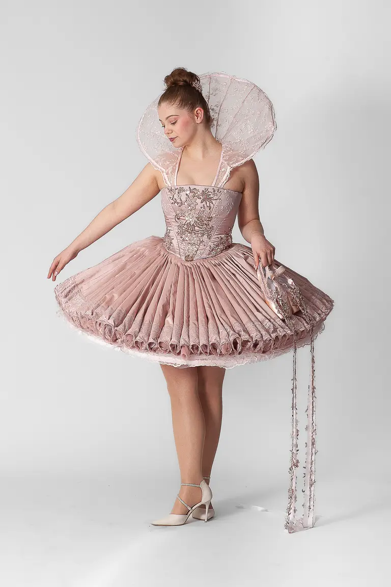 A young woman stands gracefully in a pink, voluminous dress with a fitted bodice and elaborate, ruffled details. The dress features a unique collar design and delicate embellishments. She holds a small accessory in one hand, exuding elegance against a plain white background.
