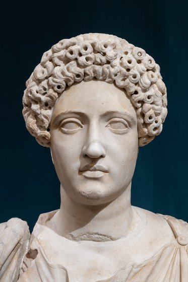 Eine antike Marmorbüste einer Person mit lockigem Haar. Das Gesicht zeigt einen neutralen Ausdruck, und die Figur trägt eine tunikaartige Gewandung. Der Hintergrund ist dunkelblau.