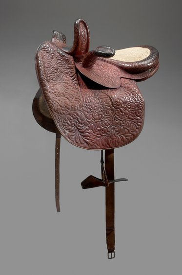 Ein dekorativer Westernsattel aus braunem Leder mit kunstvollen Mustern. Der Sattel hat eine breite Sitzfläche und hohe Sattelnähte, die auf eine robuste Verarbeitung hinweisen. Der Sattel ist auf einem grauen Hintergrund positioniert.