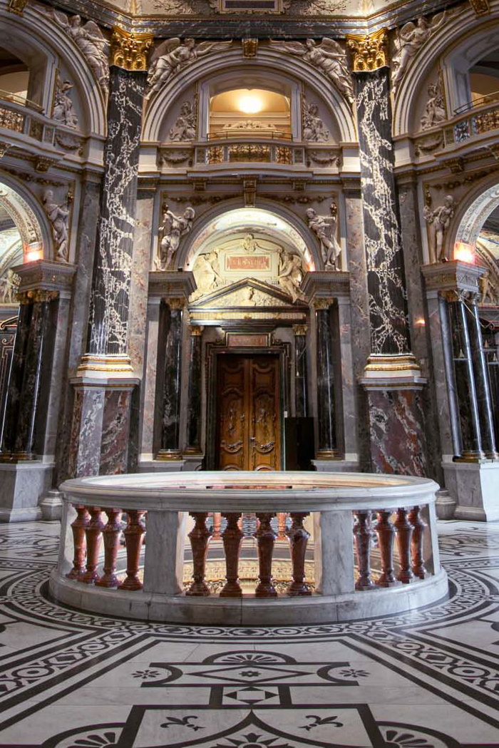 Ein eleganter Innenraum mit üppigen Marmorsäulen und kunstvollen Verzierungen. Im Vordergrund steht ein runder, marmornener Tisch mit Balustrade. An den Wänden sind detailreiche Stuckarbeiten und Bögen zu sehen. Ein großer Holztür im Hintergrund rundet die beeindruckende Architektur ab.